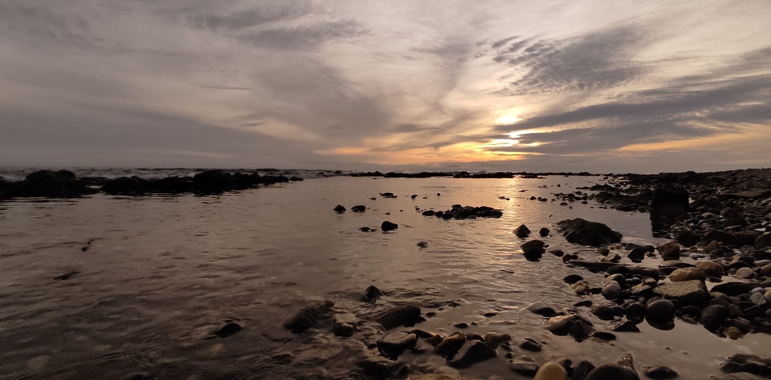 Atardecer dorado sobre una orilla rocosa y agua tranquila, con el sol reflejado en el mar, simbolizando la energía tierra del signo Capricornio