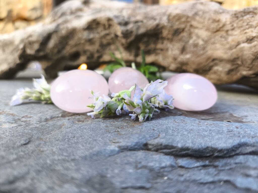 Tres piezas de cuarzo rosa pulido junto a flores de lavanda sobre una superficie de piedra, con fondo desenfocado.