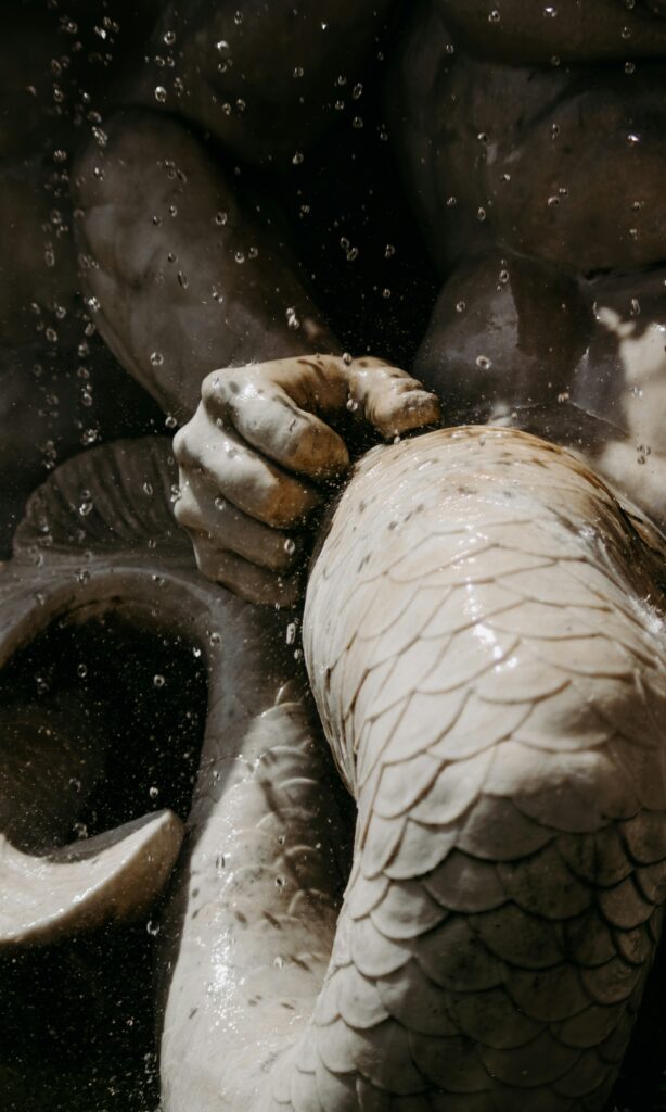 Detalle de una escultura mitológica marina en una fuente: mano de piedra y cola con escamas, con gotas de agua suspendidas en el aire.